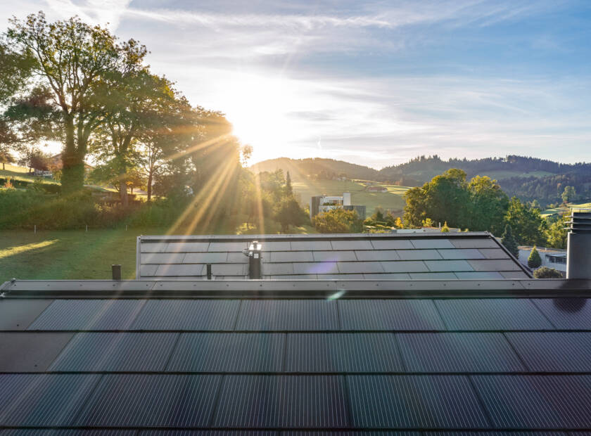 Une maison moderne avec un toit solaire. La maison est blanche avec un toit en pente et de grandes fenêtres. Le toit est recouvert d'un système de toiture photovoltaïque Sunskin Roof Lap parfaitement intégré. La maison est située sur une colline avec un grand jardin et des arbres. Maison individuelle, St. Gallen, CH